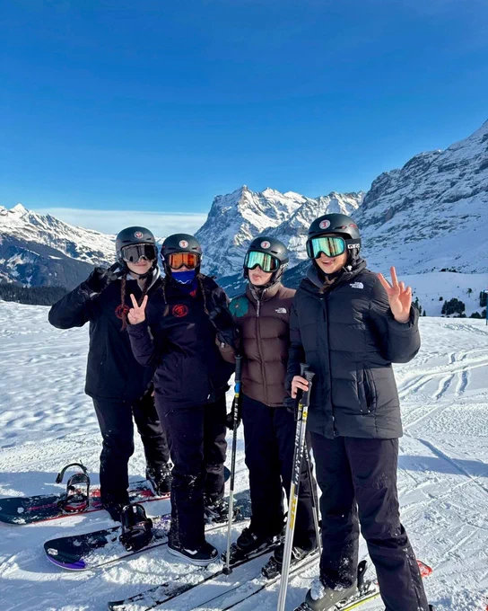 Four people are standing on a snowy mountain slope, dressed in winter gear and holding ski equipment. They are posing with peace signs. Majestic snow-covered mountains are visible in the background under a clear blue sky.