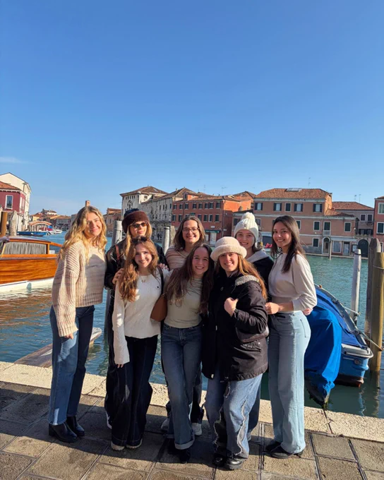 A group of people posing together on a canal walkway in Venice, with Venetian buildings and boats in the background. The sky is clear and blue.