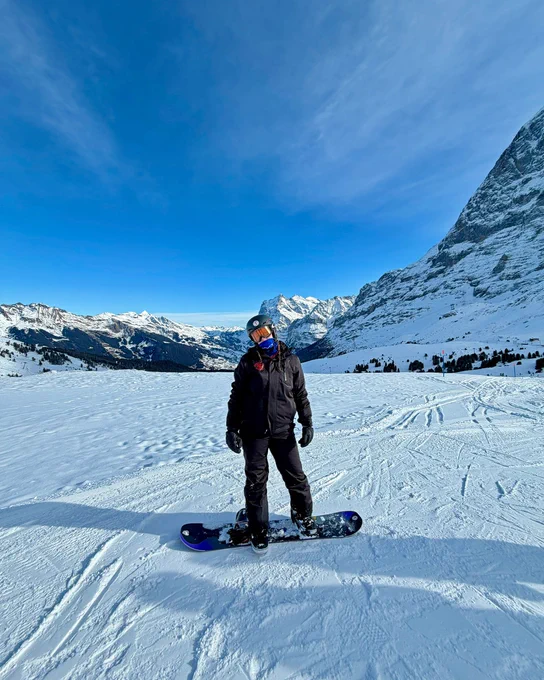 Snow-covered landscape with an individual standing on a snowboard. Snowy mountains and a clear blue sky are visible in the background, suggesting a scenic alpine setting.