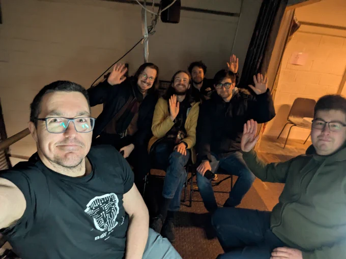 Group photo inside a workshop space: six people seated and standing, smiling and waving at the camera in a dimly lit room