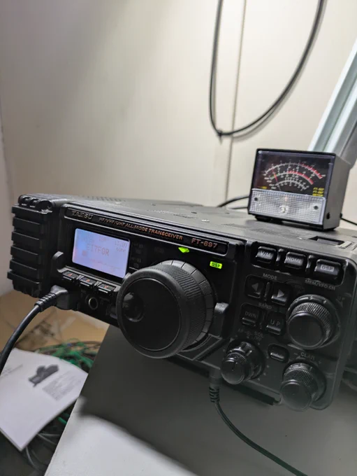 “Close-up of a Yaesu FT-897 HF/VHF/UHF all-mode transceiver on a desk with an external SWR/power meter sitting on top.”