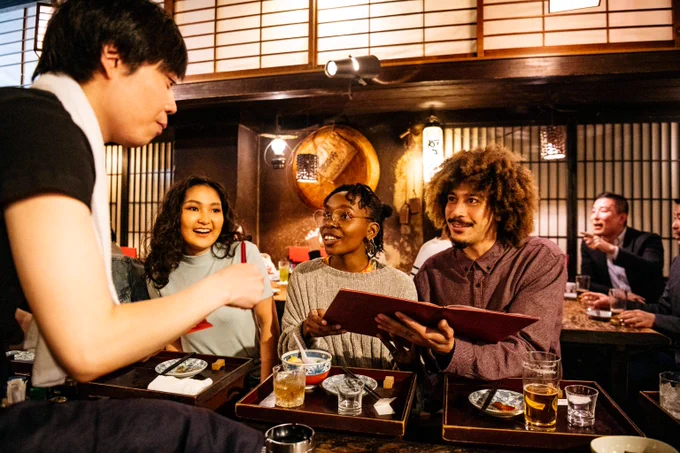 Group of friends sitting at a wooden table in a cozy Japanese restaurant, reviewing menus and enjoying drinks together.