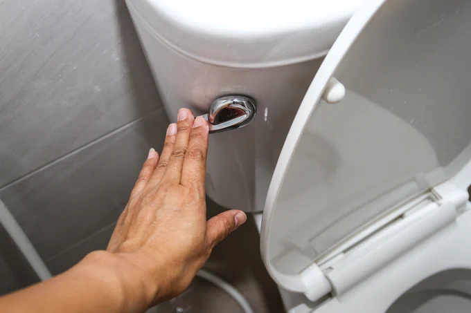 A hand is pressing the flush button on a toilet in a bathroom. The toilet seat lid is partially open.