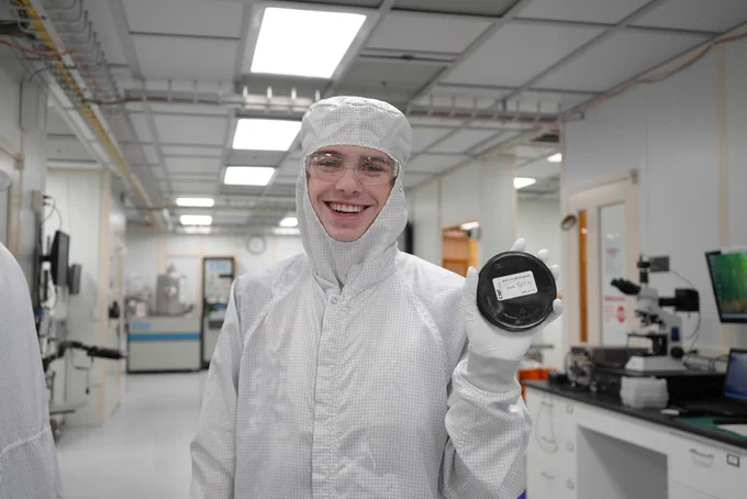 A person in a cleanroom suit holds up a silicon wafer inside CNF laboratory. Scientific equipment and computers are visible in the background.