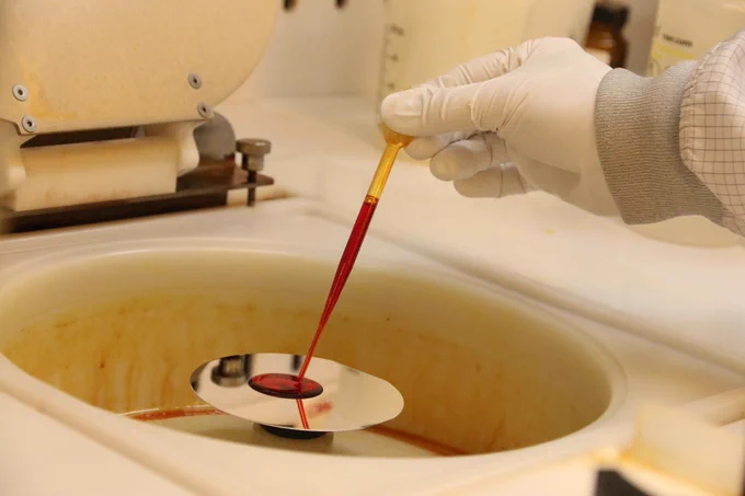 A gloved hand holding a pipette is applying a red solution onto a small, reflective silicon wafer in a laboratory setting.