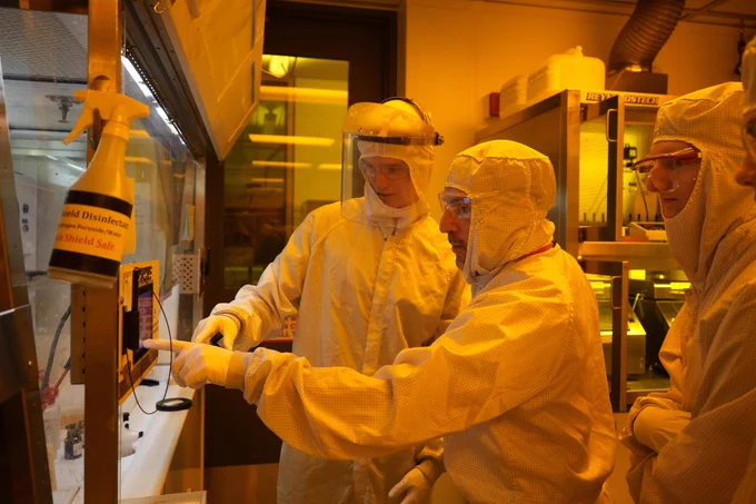 Laboratory workers in cleanroom suits and protective gear are gathered around a control panel in a laboratory. The environment is illuminated with an amber light. One individual is pointing at the panel, while others observe. Various lab equipment is visible in the background.