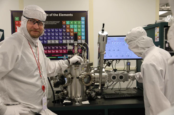 Individuals in cleanroom suits work with scientific equipment in a lab. A Periodic Table of the Elements is visible on the wall behind them.