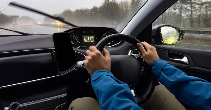 View from inside a car showing a person's hands on the steering wheel, driving on a rainy day with other vehicles visible ahead on the highway. The dashboard displays the car's speed and temperature.
