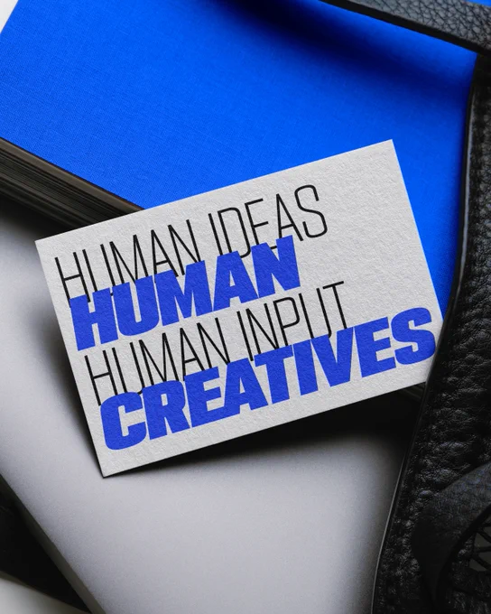 A card placed on a blue and gray surface features bold black and blue text with the phrases "Human Ideas," "Human Input," and "Human Creatives." The background includes parts of a textured black material.