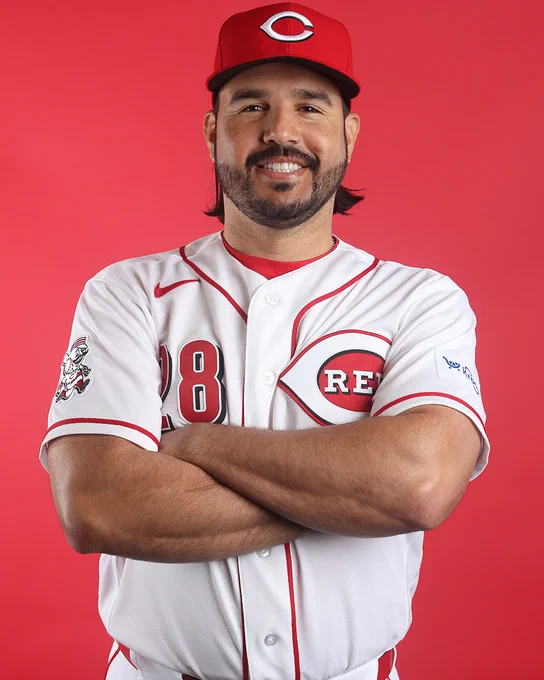 Eugenio Suárez smiles for a picture with his arms crossed while wearing a home white Cincinnati Reds uniform and red Cincinnati Reds cap.