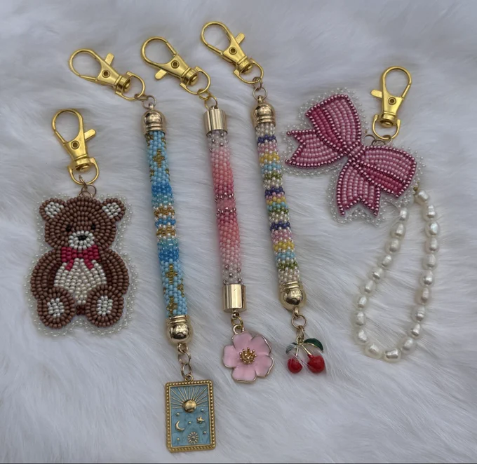 A ssortment of beaded bag charms laying on white rabbit fur. 3 peyote stitch tube shaped with enamel charms on end - one pink with flower charm, one blue with tarot card charm, one cream and pastel rainbow with a cherry charm. Flat stitched charms in teddy bear design and bow design with string of freshwater pearls.
