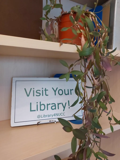 Small Visit Your Library sign noting @Library4NUCC handle on a shelf behind a flowing green plant