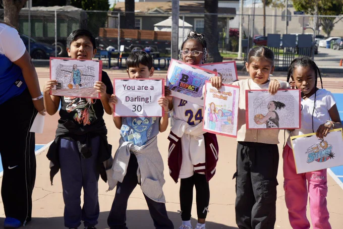 Five kids hold drawings outside. The children stand on a blue and red surface, showcasing their artwork. One drawing shows a basketball player.