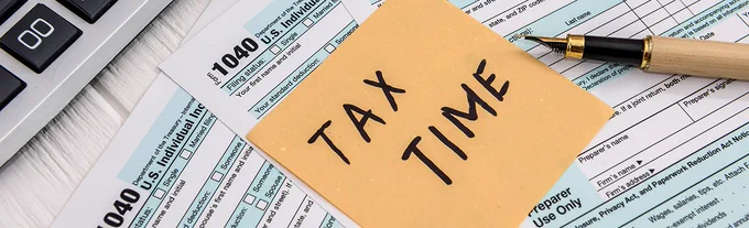 Tax documents on a table with a note on the front that reads "Tax Time". A pen is resting at the right.