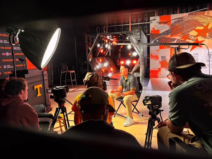 A photograph of a man sitting in a chair wearing an orange shirt with the logo T behind him in orange. He is being interviewed and there are cameras in front of him and lights. The background has a multicolored orange wall and orange and white lights.