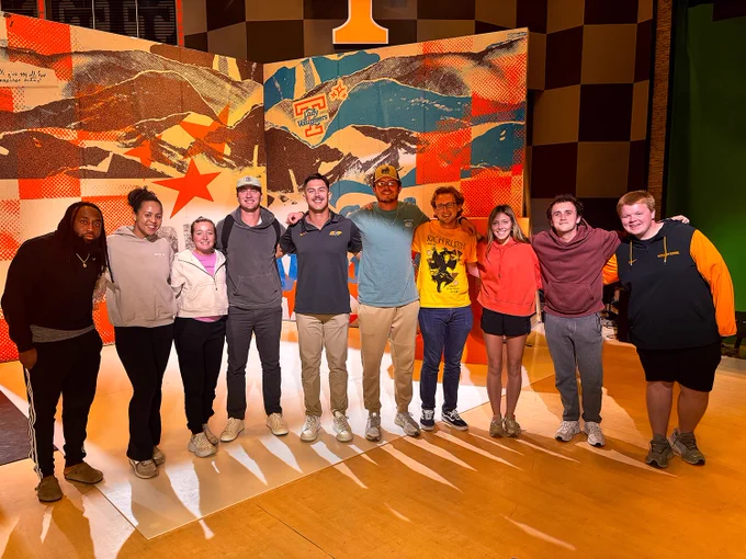Group photo of students pose for a photograph with Vols baseball player Hunter Ensley in a orange lit room with a multicolored background.
