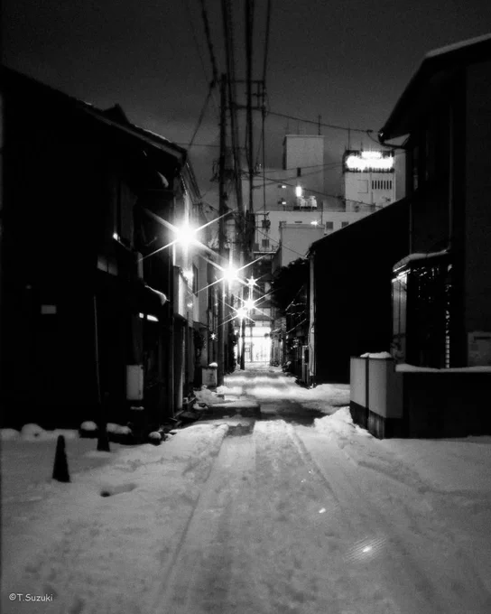 雪の残る夜の路地。街灯がにじみ、奥の明かりへ道が伸びている。
A snowy alley at night; streetlights glow softly as the path leads to a distant light.