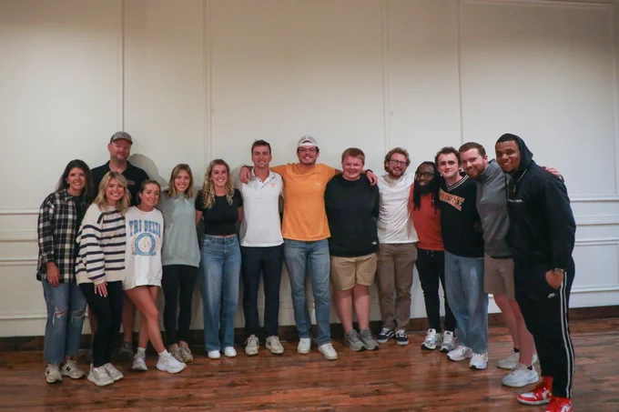 A group of students and faculty members pose for a photograph with a white wall behind them.