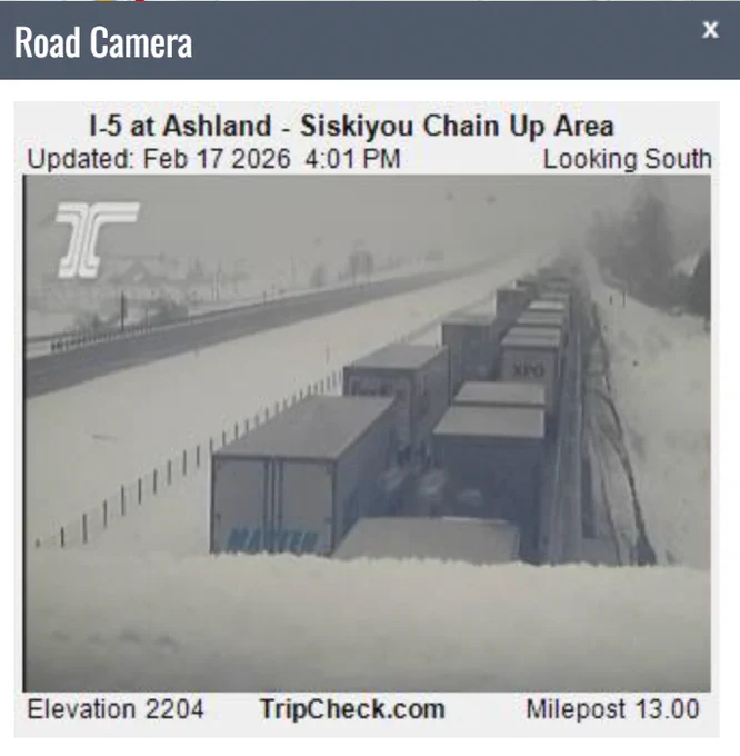 A TripCheck camera view of I-5 traffic waiting during a hold at the Oregon to California border while crews plow the highway. This view looks south and shows heavy snow in the area.