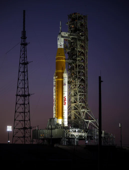 NASA\u2019s Artemis II Space Launch System (SLS) rocket and Orion spacecraft are seen illuminated by lights at Launch Complex 39B, Tuesday, Feb. 10, 2026, at NASA\u2019s Kennedy Space Center in Florida. The orange core stage stands between two white solid rocket boosters and is topped with the white Orion spacecraft. The trussed mobile launcher structure stands behind the rocket and spacecraft. A lightning tower can be seen to the left of the rocket. The sky in the background is a dusty purple hue. Credit: NASA/Aubrey Gemignani