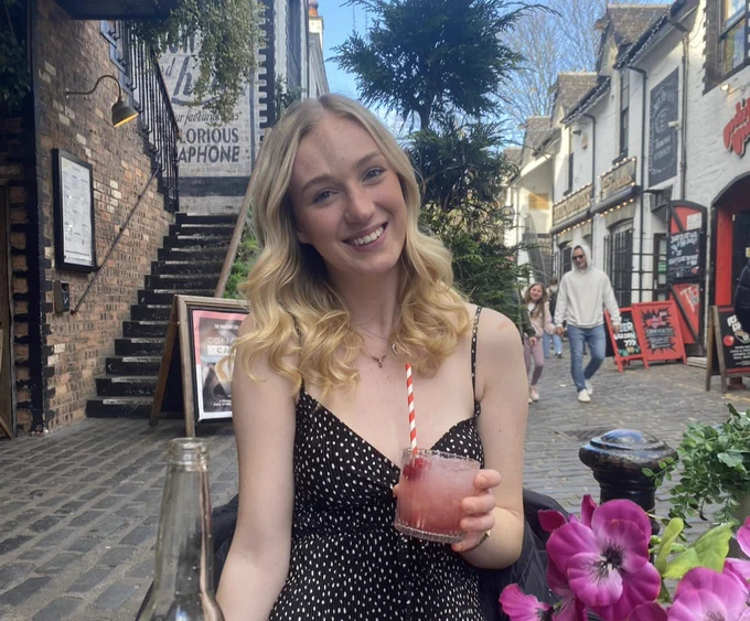 Holly is a white woman in her early twenties with long blonde hair. She has a "sabre strike" indentation in a vertical line on her forehead. She sits in an outdoors restaurant sipping on a red cocktail, smiling at the camera.