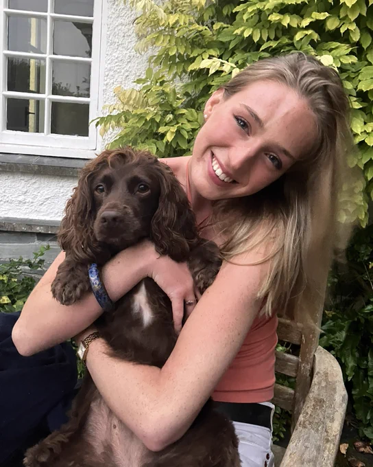 Holly has her brown cocker spaniel on her lap and smiles at the camera. The spaniel sits on his hind legs so you can see his belly and white patch on his chest.