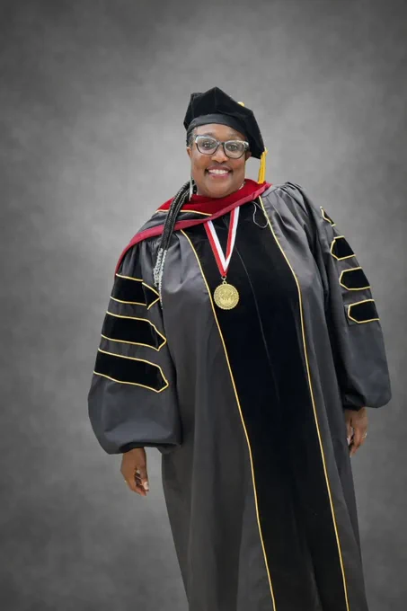 Linda Davis wearing a charcoal gray commencement robe with black and gold striped adornments, red sashes, a black cap with a yellow tassle, and golden medal with a red and white ribbon. Linda is a young Black woman smiling vibrantly, her long beaded braids flowing over her right shoulder.