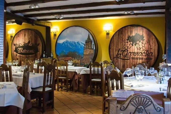 Interior de un restaurante de estilo rústico, con paredes en tonos amarillos y vigas de madera en el techo. Las mesas están vestidas con manteles blancos y marrones, preparadas con copas de vino y cubertería cuidadosamente colocada.

En la pared del fondo destacan tres grandes toneles decorativos de madera. En uno se lee “Malvasía” y en otro “La Garnacha restaurante asador”, mientras que el central muestra un paisaje montañoso con una torre histórica. El ambiente combina tradición y calidez, evocando gastronomía local y una experiencia acogedora en un entorno cuidado.
