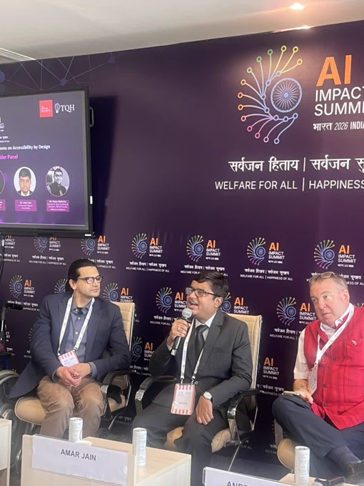 Amar Jain speaking into a handheld microphone during a panel at the AI Impact Summit 2026. He is seated between two fellow panelists against a purple AI Impact Summit backdrop that reads “Welfare for All | Happiness for All.” A screen beside them displays session details and speaker photos.