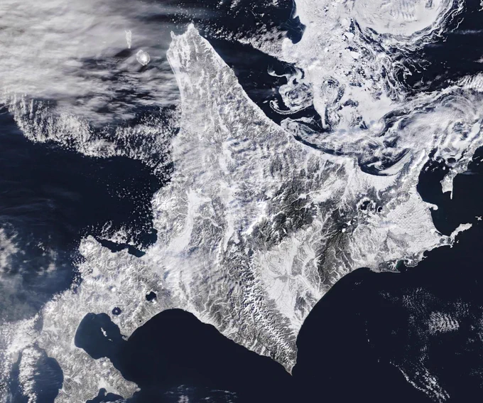 A satellite image of snow-covered landscapes across Hokkaido. With more than 31 active volcanoes, the island features several large caldera lakes, including at least five that are visible in the image. In the east, forested windbreaks around Nakashibetsu form a checkerboard pattern, while to the north, swirls of drifting sea ice adorn the Sea of Okhotsk.