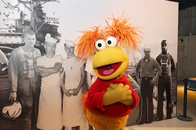 A furry, orange puppet with wild hair stands in front of a black and white photo of three sailors and two Navy officers on a ship.