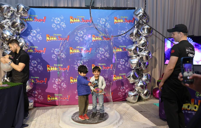 Young children standing inside a large bubble display at a Gazillion Bubble Show event, with metallic balloon decorations and a branded backdrop. An onlooker captures the moment with a mobile phone.