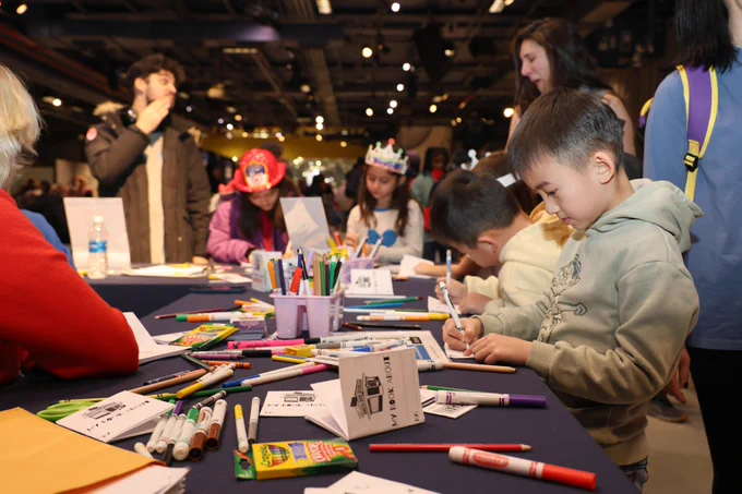 A group of children are engaged in an arts and crafts activity at a table filled with markers, crayons, and paper. The room is well-lit and bustling with people, suggesting a lively event.