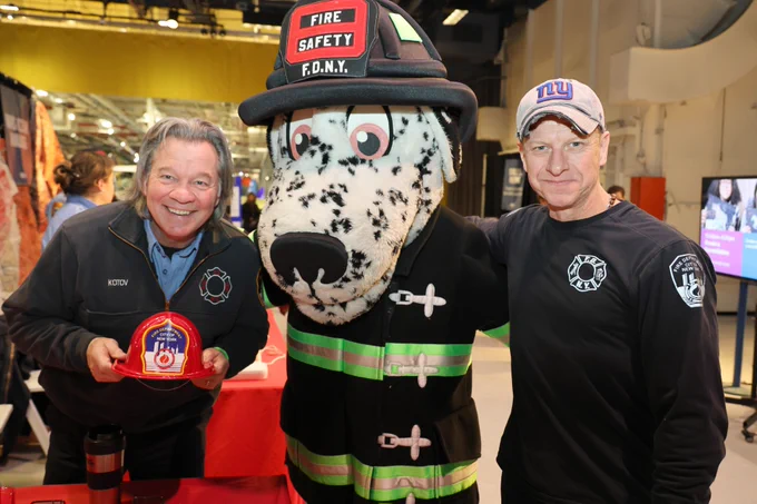 Two individuals are posing with a large Dalmatian mascot dressed in a firefighter outfit labeled "F.D.N.Y." They are at a booth featuring fire safety materials. One person is holding a small, red firefighter helmet. The other is wearing a New York Giants cap.