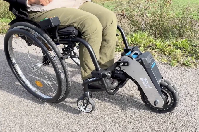 Side view of a manual wheelchair fitted with a WheelMove powered front-wheel attachment, showing the motorized wheel, battery pack, and mounting arms extending from the footrest area as the user sits on a paved path outdoors.