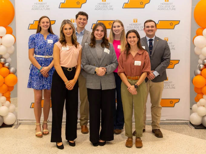 A group of seven students stands in front of a backdrop with the Herbert College of Agriculture logo. They are dressed in business-casual attire. Balloons in white, orange, and silver are on either side.