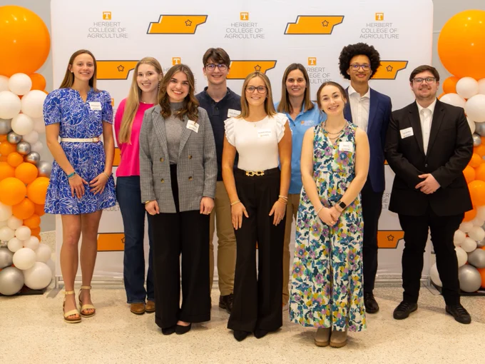 A group of eight students and one professor standing together in front of a "Herbert College of Agriculture" backdrop, surrounded by orange, white, and silver balloons. All are dressed in business-casual attire.