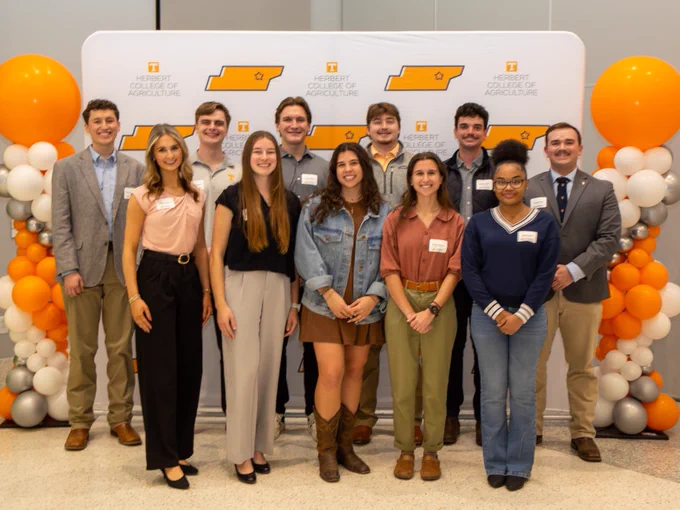A group of eleven students standing in front of a "Herbert College of Agriculture" backdrop and orange, white, and silver balloons on both sides. They are dressed in business-casual attire, smiling at the camera.