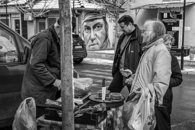 Black and white documentary photograph of people buying smoked meat from a street vendor on Badnji Dan in Zvecan. A mural portrait looms in the background, adding cultural and social context. Photograph by Serbian photographer from Kosovo and Metohija, Ivan Maksimovic.