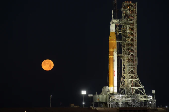 NASA's Space Launch System rocket, an orange cylinder with twin white boosters, stands on the launch pad at NASA's Kennedy Space Center. A full, orange-red Moon hangs low in the dark night sky; the rocket is brightly lit from the ground. Credit: NASA/Ben Smegelsky