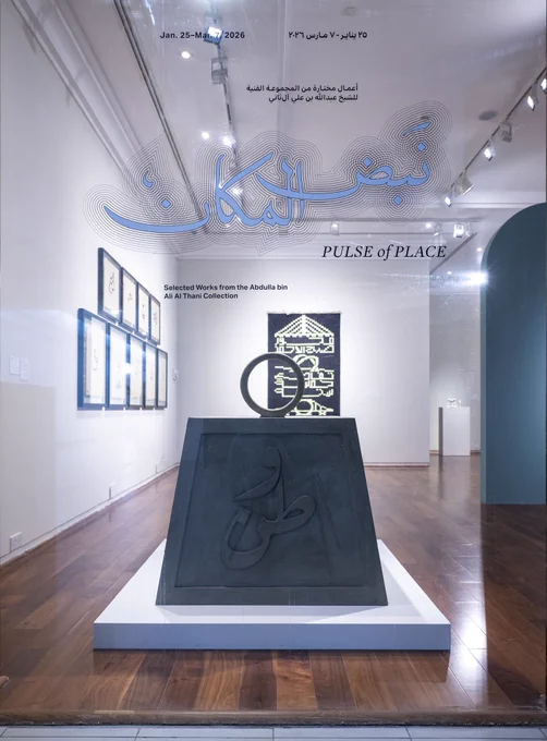 This image shows the entrance to an art exhibition titled "PULSE of PLACE." The view is through a glass window or door, with text and graphics layered over a brightly lit gallery space.
Key Visuals
• Text & Graphics: Large, stylized light-blue Arabic calligraphy spans the center of the glass, surrounded by thin concentric outlines. Below it, the English title "PULSE of PLACE" appears in a serif font. Smaller text indicates the dates (Jan. 25–Mar. 7, 2026) and notes that these are "Selected Works from the Abdulla bin Ali Al Thani Collection."
• Central Artwork: Directly inside, a large, dark grey trapezoidal sculpture sits on a white rectangular base. It features raised Arabic calligraphy on its face and is topped with a circular metal ring.
• Gallery Interior: The room has polished wood floors and white walls. To the left, several framed artworks are hung in a neat grid. In the background, a dark tapestry with geometric yellow designs is visible.