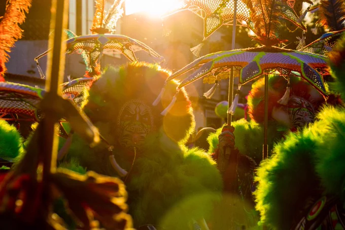 A imagem retrata bate-bolas, figuras tradicionais do Carnaval carioca, captados em cena de forte impacto visual e simbólico. Os personagens aparecem parcialmente envoltos por sombras criadas pelo contraste com a luz dourada do sol ao fundo. Essa luz atravessa a cena por trás dos personagens e cria silhuetas marcadas, ressaltando as texturas dos adereços. Os bate-bolas seguram sombrinhas decoradas. As fantasias são volumosas e chamativas, com destaque para o uso abundante de plumas e materiais felpudos em tons vibrantes. As máscaras, parcialmente visíveis, reforçam o anonimato dos foliões.