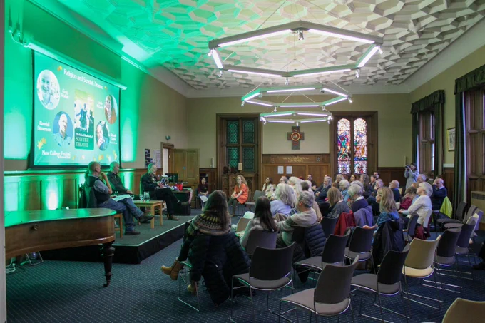 Colour photo of people sat in the Martin Hall looking at four speakers sat on the stage in the hall