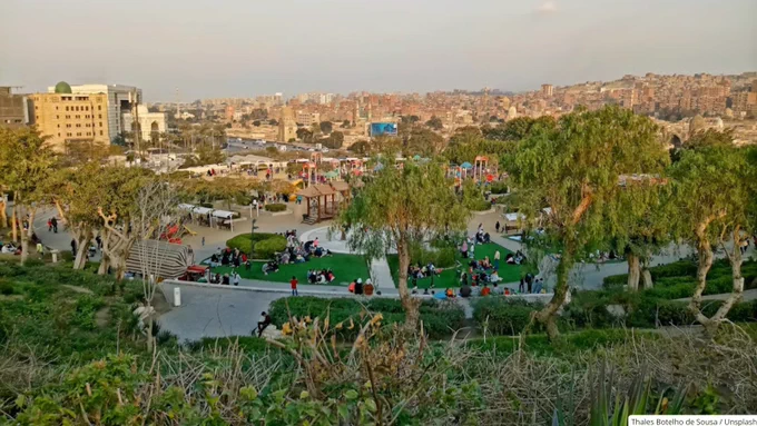 A lively urban park with groups of people relaxing on green lawns surrounded by trees and pathways under a clear sky.