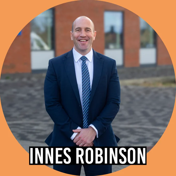 Portrait of Innes Robinson wearing a navy suit and striped tie, standing outdoors in front of a brick building. The image is framed in an orange circle with the name “Innes Robinson” displayed at the bottom.