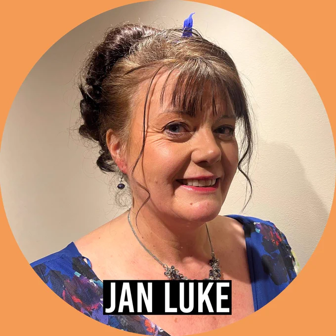 Portrait of Jan Luke wearing a blue floral dress and necklace, smiling against a neutral indoor background. The image is framed in an orange circle with the name “Jan Luke” displayed at the bottom.