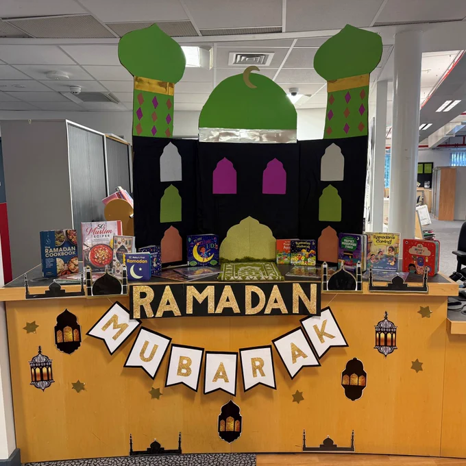 A Ramadan display on a library desk featuring Ramadan themed cookbooks and picture books. On the desk there is a model of a mosque made out of paper. The banner hanging from the desk reads 'Ramadan Mubarak'