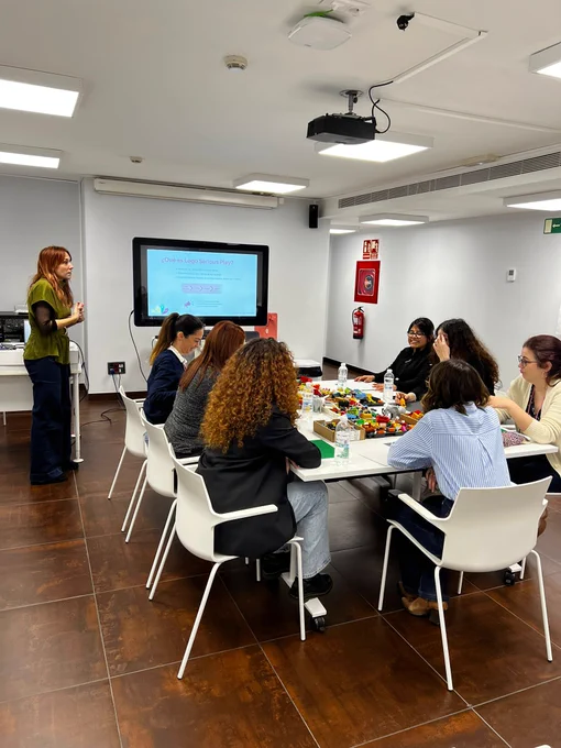 Al fondo se ve una pantalla mostrando la presentacion sobre LSP. En un lateral a la formadora dando instrucciones. Y la mayor parte de la imagen es la mesa al rededor de la cual se sientan 7 alumnas con piezas de lego.
