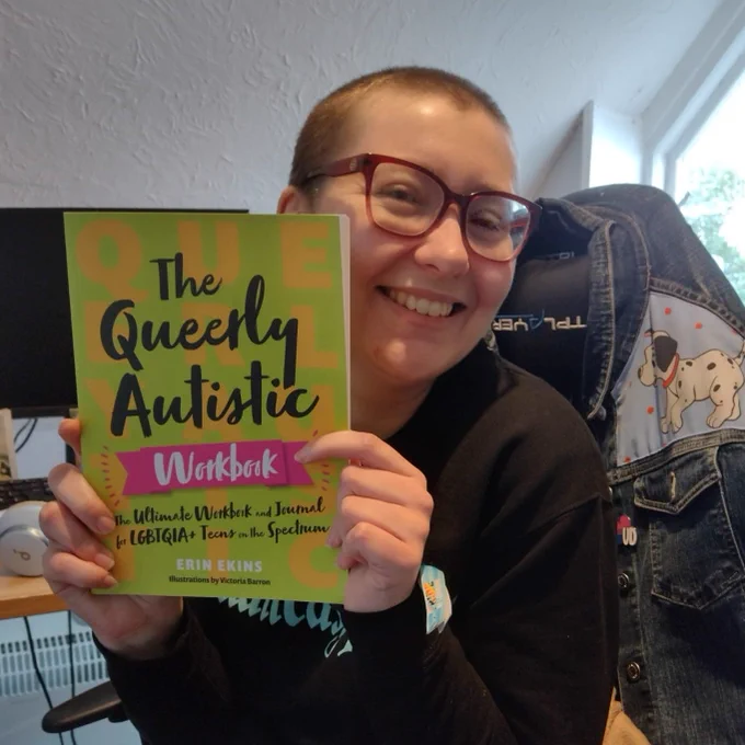 Me, a white woman in my mid 30s with a buzzcut and glasses, grinning and holding up a copy of The Queerly Autistic Workbook: The Ultimate Workbook and Journal for LGBTQIA  Teens on the Spectrum. 