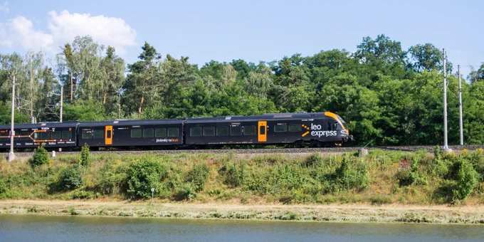 A Leo Express train travels on tracks adjacent to a river, surrounded by greenery and under a clear blue sky.
